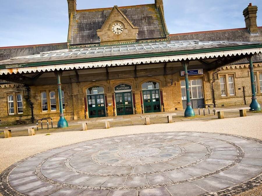 A historic train station with a clock tower, a covered waiting area, and a circular stone pavement in the foreground.