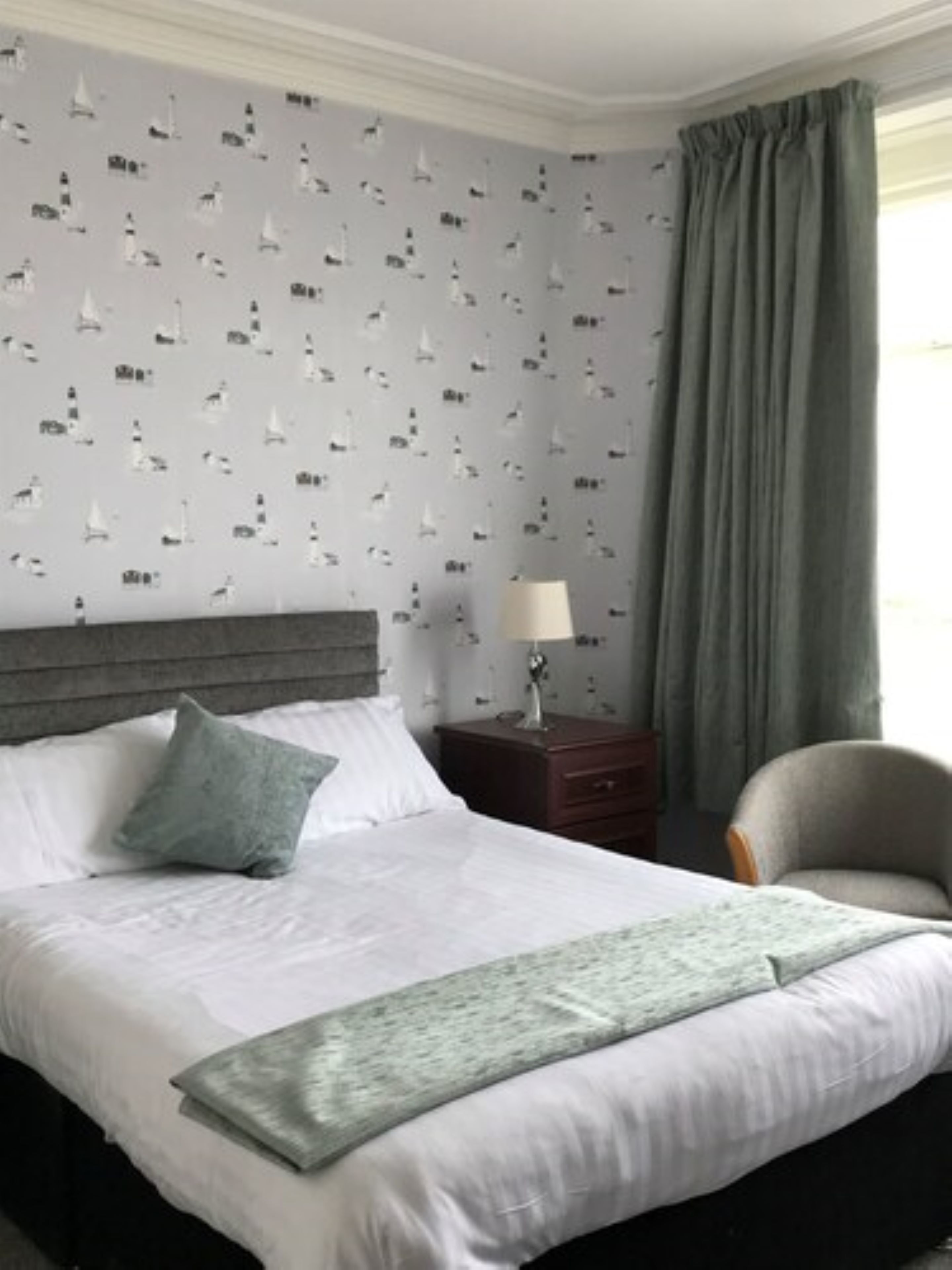 A neatly made bed with white linens and a gray throw blanket, a gray upholstered chair, a wooden nightstand with a lamp, and a wall with a nautical-themed wallpaper.