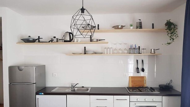A modern kitchen with white cabinets, a stainless steel refrigerator, a gas stove, and a built-in oven. There are wooden shelves above the counter holding various kitchen items. - Anica Apartmani (Funtana)