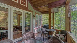 A covered outdoor deck with wicker furniture, glass doors leading to an adjacent patio, and a view of the surrounding forest. - A Foxfire Retreat: Gated Resort w/ Pools & Hot Tub, Fishing, Fitness Area (Boone)
