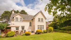Une maison à deux étages avec une pelouse bien entretenue, une terrasse dotée de mobilier d’extérieur et une variété d’arbustes et d’arbres.