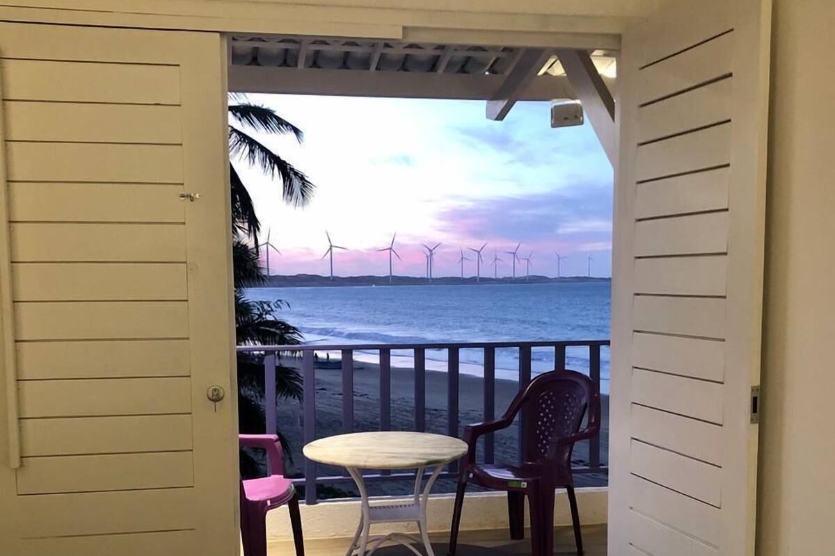 A room with a view of the ocean and wind turbines, featuring a bed with a fish pattern bedspread, a small round table, and two chairs.