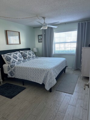 Une chambre à coucher comprenant un lit, un ventilateur de plafond, une fenêtre avec des rideaux, une lampe de chevet et une commode.