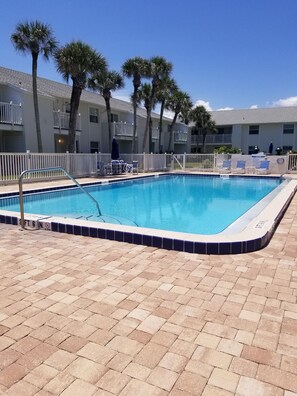 Une piscine rectangulaire dont le bord est recouvert de carreaux bleus, entourée d’une terrasse en briques. On aperçoit en arrière-plan des clôtures blanches et des palmiers.