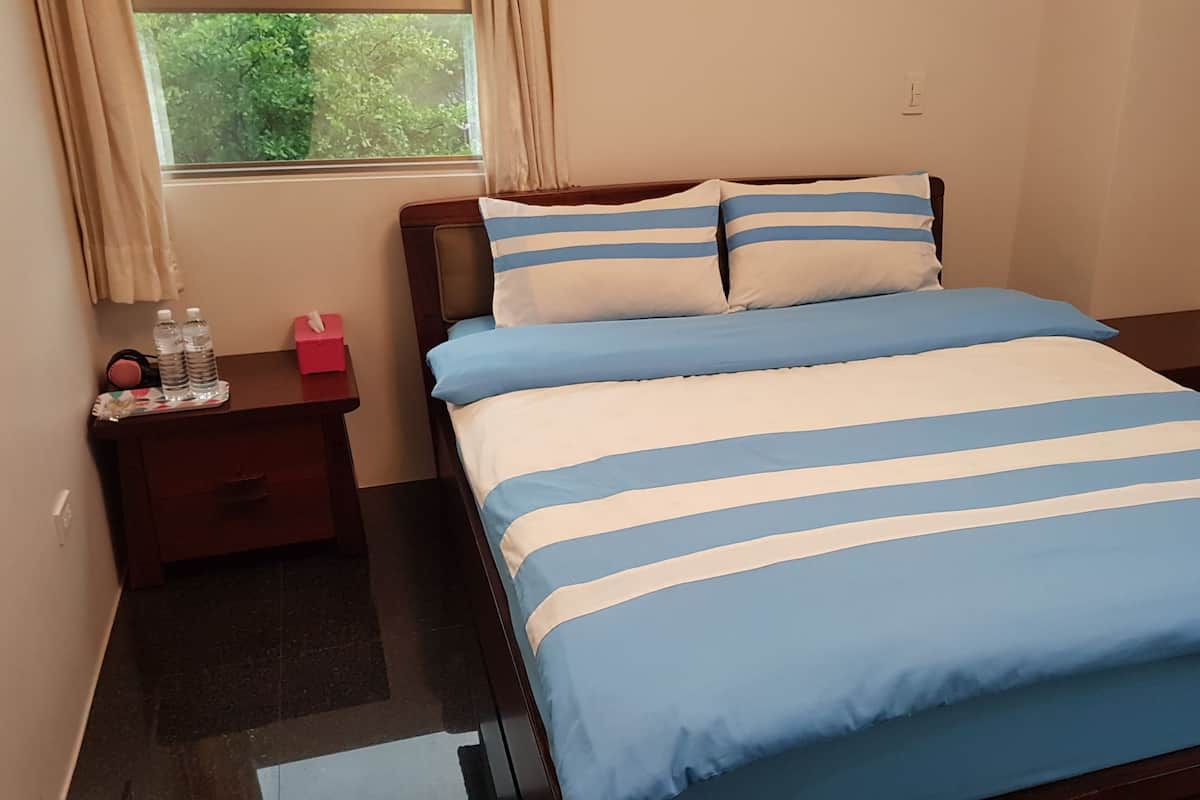 A bedroom with a bed, two nightstands, a window with curtains, and a small table with water bottles and a tissue box.