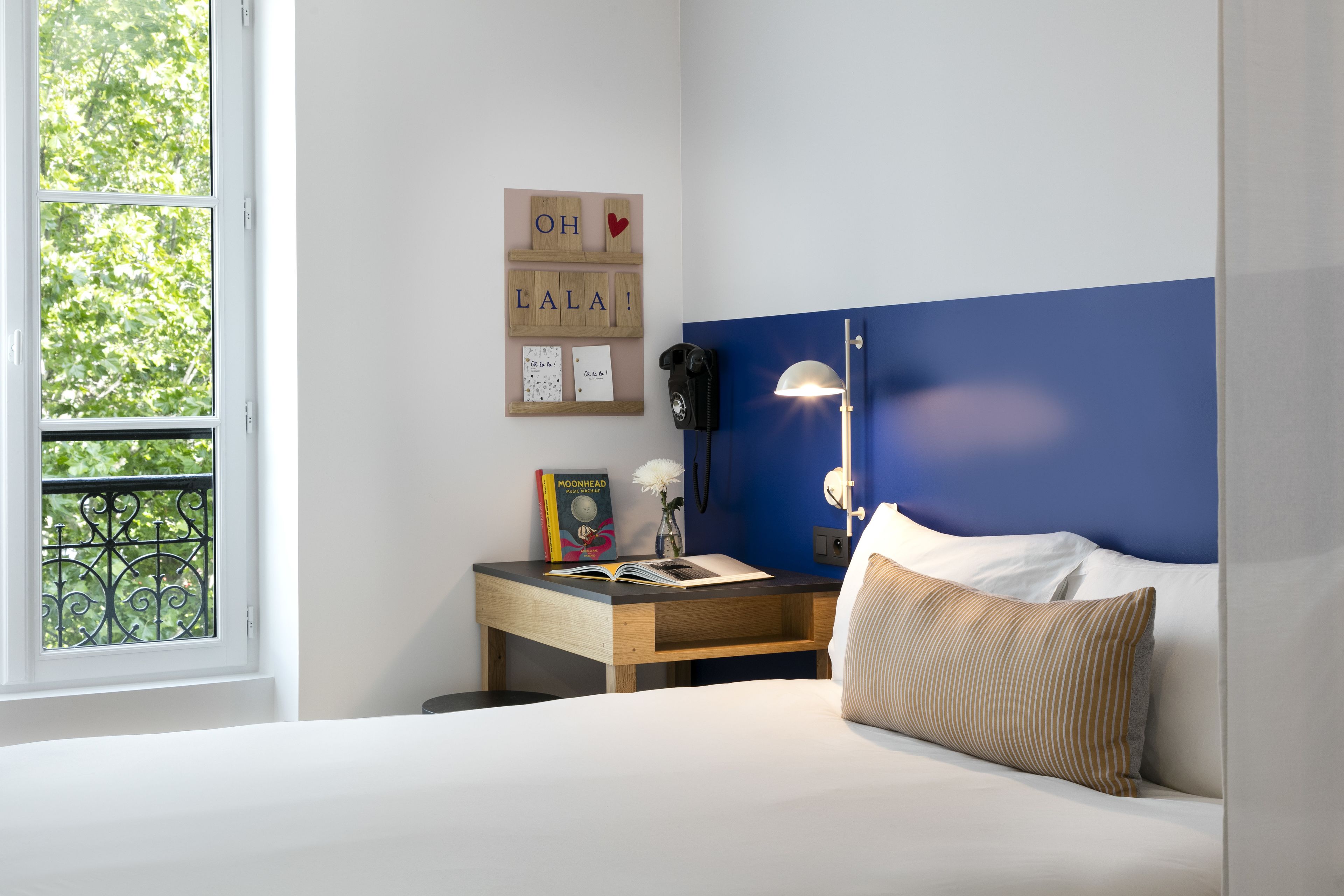 Un dormitorio con una cama, una mesita de noche con una lámpara, una mesita con un libro y un florero, y una ventana con vistas a árboles.