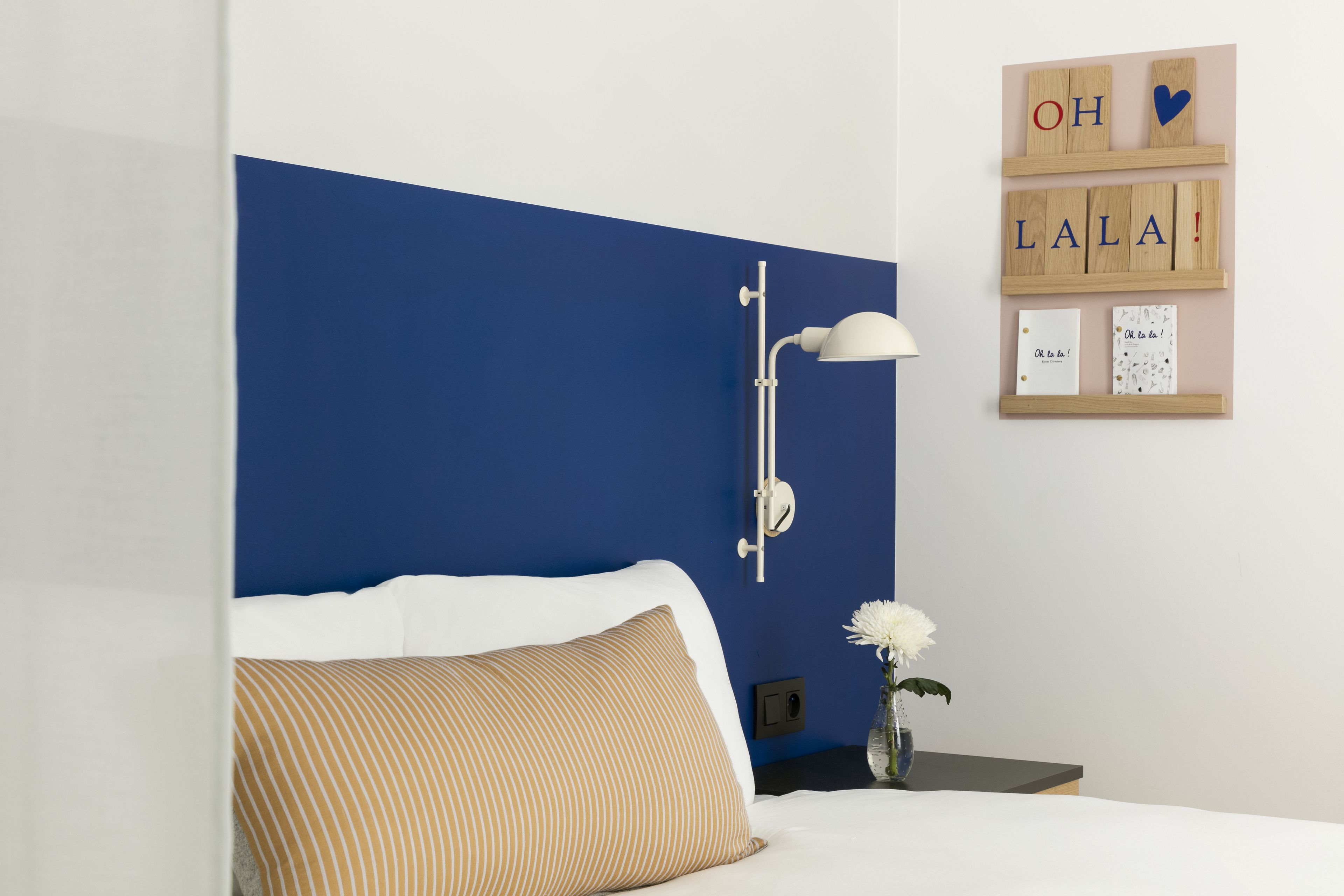 A bedroom with a blue accent wall, a white bed with a striped pillow, a bedside table with a vase of flowers, and a wall-mounted lamp.