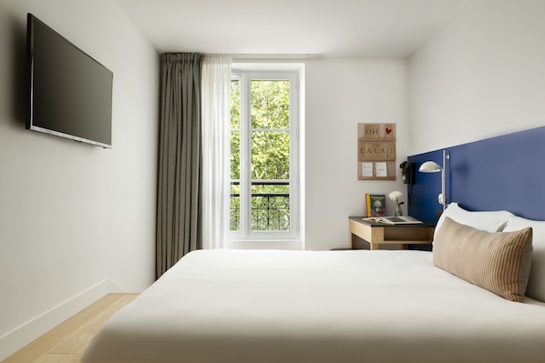 Ein Schlafzimmer mit einem großen Bett, einem Flachbildfernseher, einem Fenster mit Vorhängen, einem Nachttisch mit Büchern und einer Lampe sowie einer blauen Akzentwand.