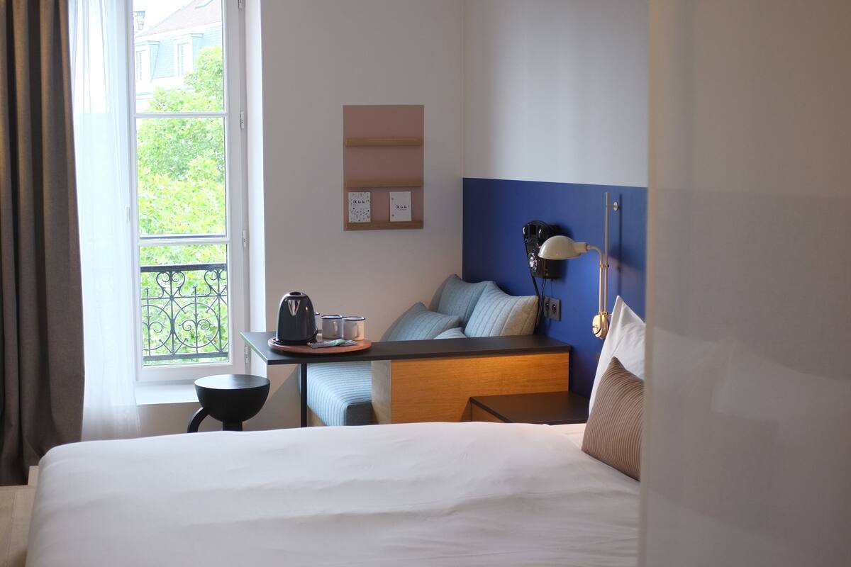 Habitación de hotel con una cama grande, un escritorio con hervidor, una lámpara y vistas a zonas verdes al exterior.