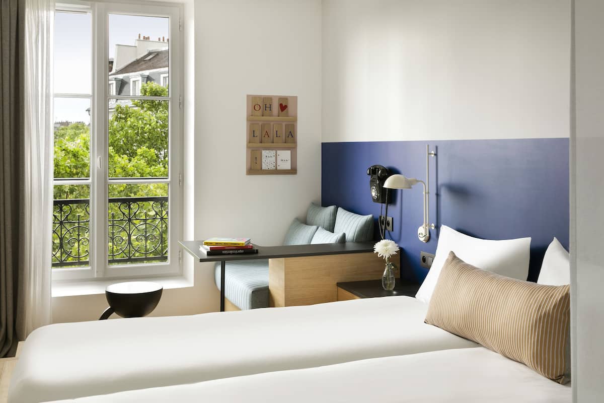 Habitación de hotel con cama, escritorio, silla, ventana con vista a follaje, y pared con detalle azul y un tablero con letras y símbolos.