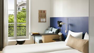 Habitación de hotel con cama, escritorio, silla, ventana con vista a follaje, y pared con detalle azul y un tablero con letras y símbolos.
