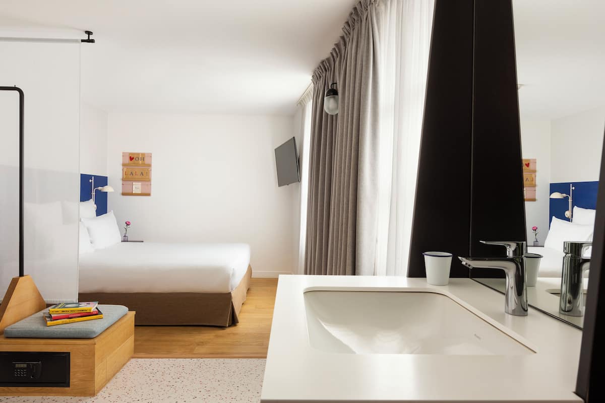 Una habitación de hotel moderna con una cama, un lavabo blanco, un espejo y un televisor en la pared.