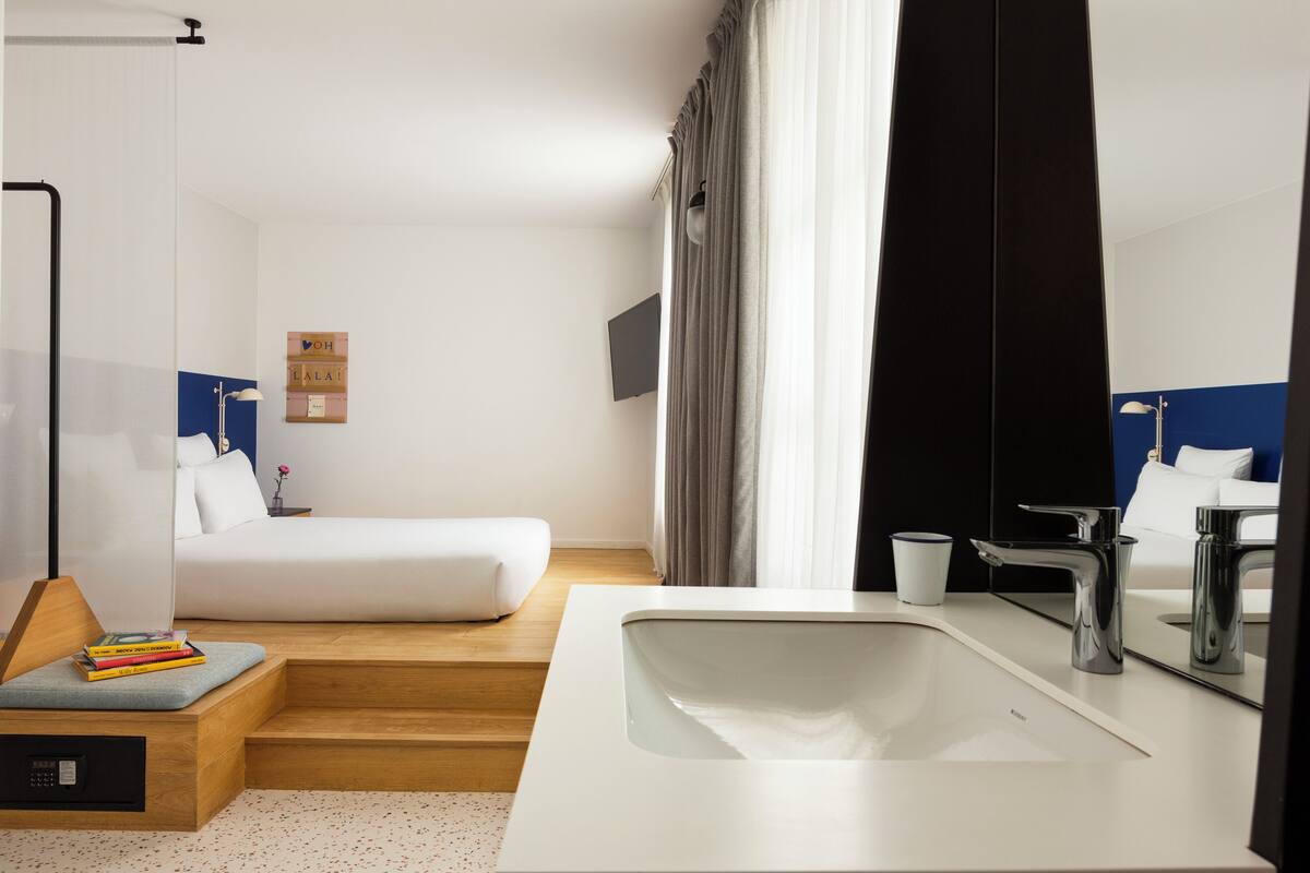 Una habitación de hotel con cama, mesita de noche con libros, lavabo blanco y espejo.