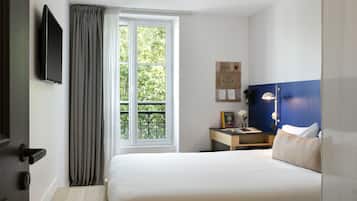 A hotel room with a bed, a bedside table, a lamp, a TV mounted on the wall, and a window with curtains.