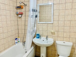 A bathroom with a bathtub, a sink, and a toilet. There is a shower curtain, a mirror, and a shelf with toiletries.