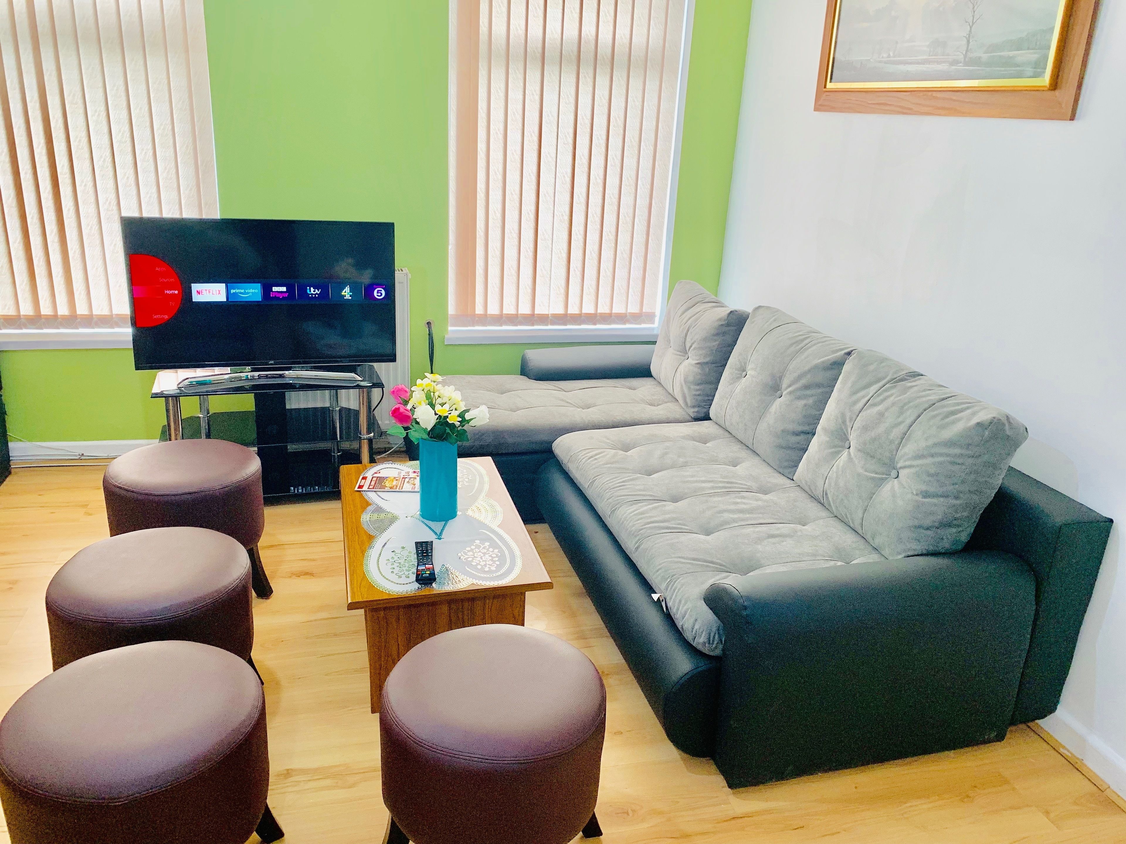 A living room with a grey sofa, two brown ottomans, a coffee table with a vase of flowers, a flat-screen TV, and a small table with a remote control.