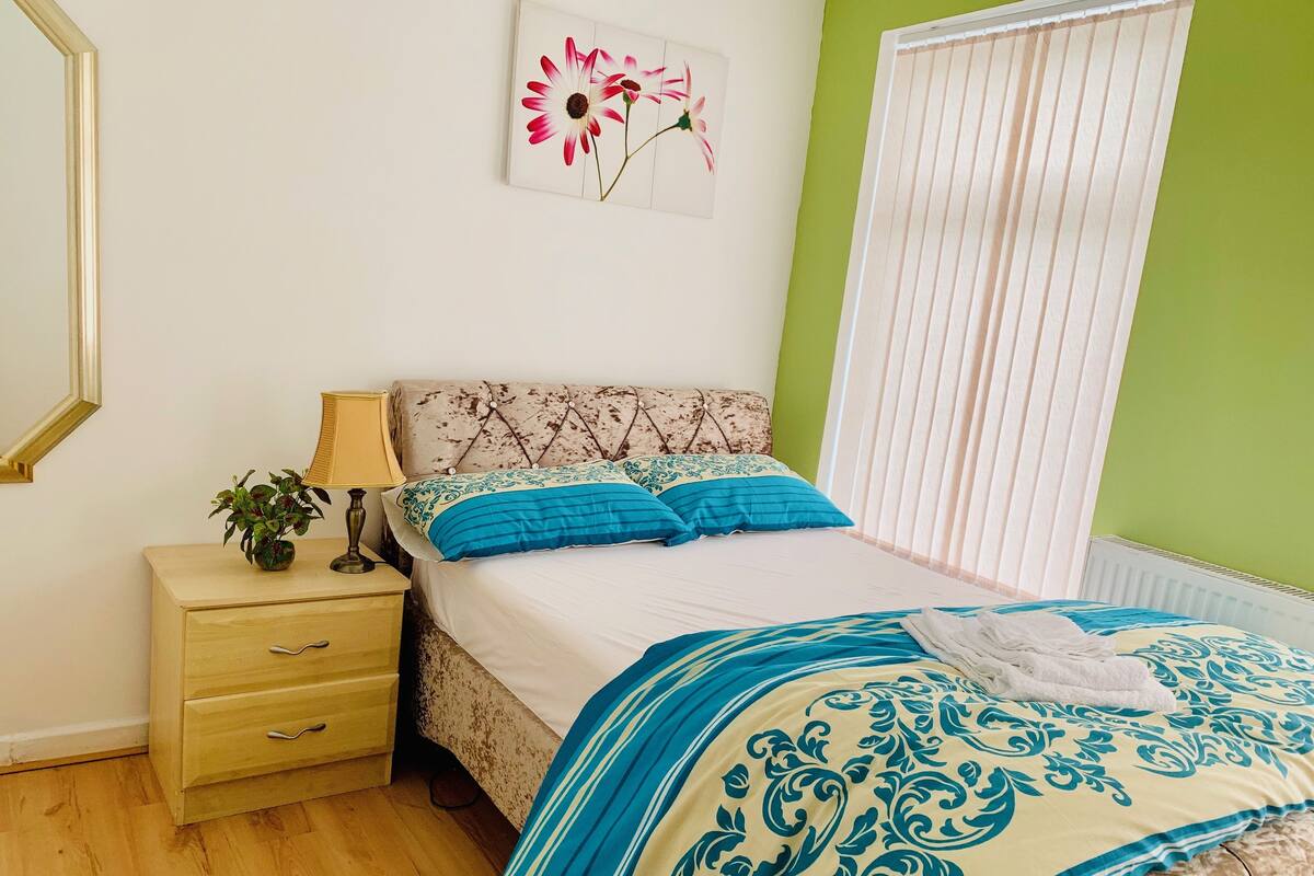 A bedroom with a bed, a nightstand, a lamp, a framed picture on the wall, and a window with blinds.
