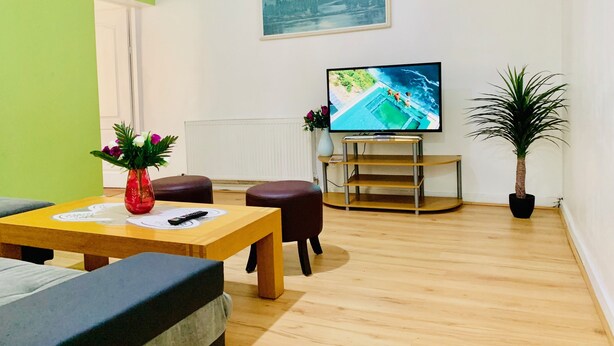 Una sala de estar con una mesa de madera, dos otomanas de cuero marrón, un televisor sobre un mueble bajo, una planta en maceta y un cuadro en la pared.
