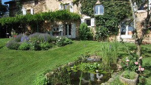 A stone building with ivy-covered walls, a small pond with a statue, and a well-maintained garden with various plants and flowers. - La Basserie (Laguepie)