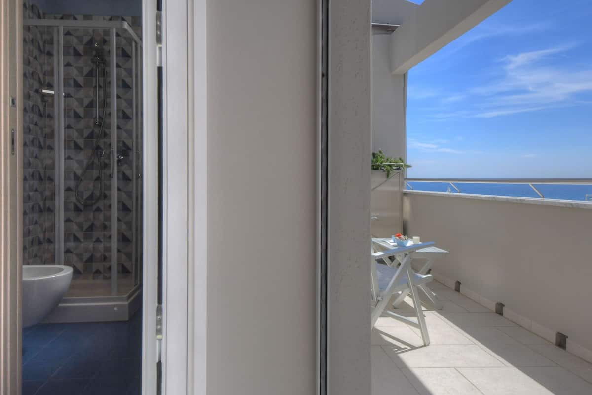 A balcony with a view of the sea, a glass-enclosed shower, and a small table with chairs.