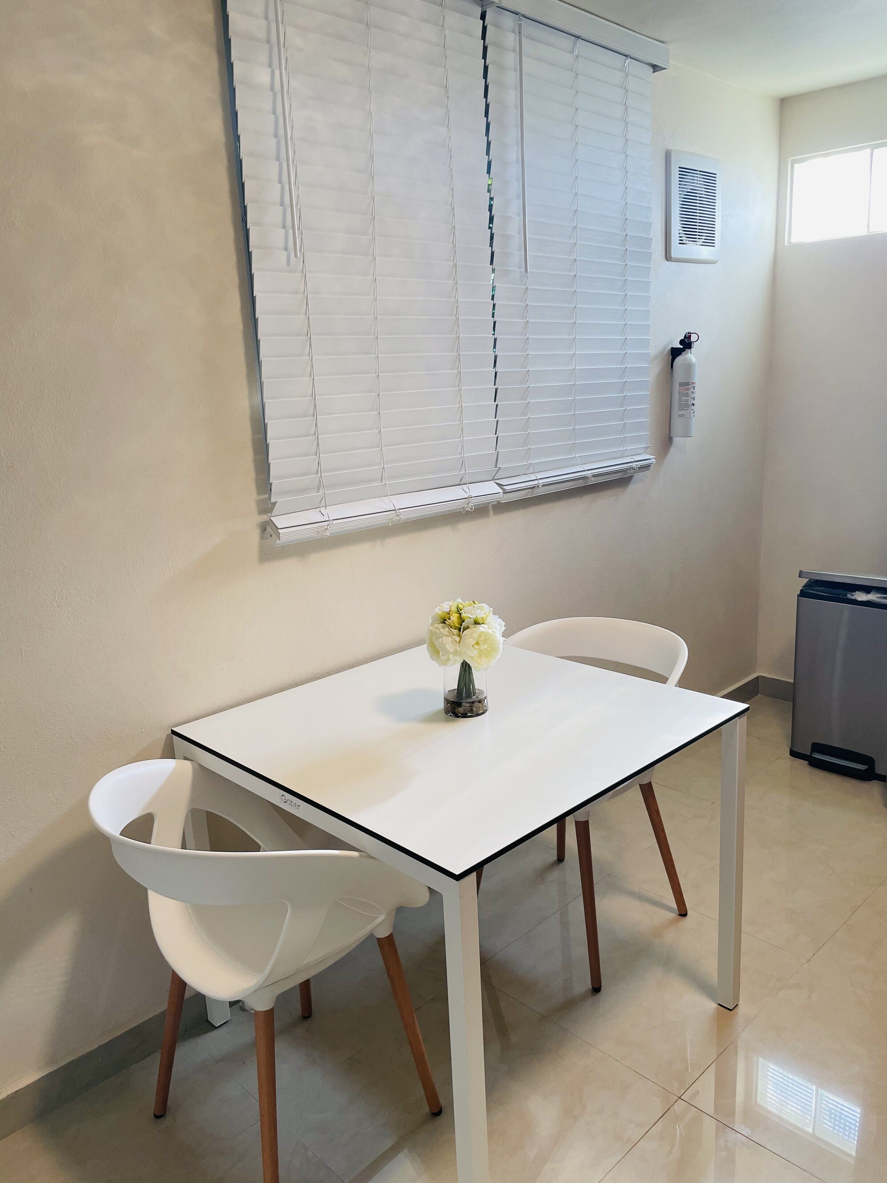 Una mesa blanca con dos sillas, un florero con flores, una ventana con persianas, un extintor y un cubo de basura.