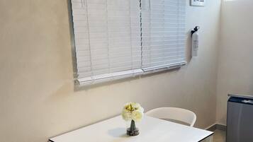 Una mesa blanca con dos sillas, un florero con flores, una ventana con persianas, un extintor y un cubo de basura.