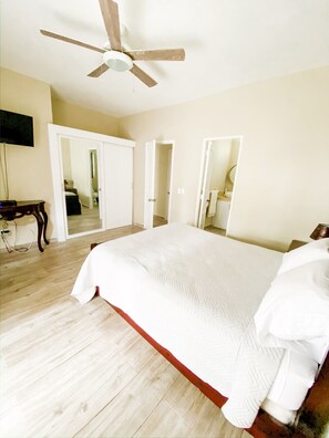 A bedroom with a large bed, wooden floor, ceiling fan, and a TV on a stand.