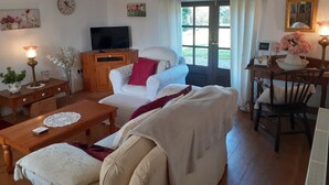 A cozy living room with a white sofa, wooden coffee table, and a television on a wooden cabinet. A window with curtains is visible, and a small table with a lamp and flowers is on the right. - 300 Year old Tradition Irish Cottage (Rathkeale)