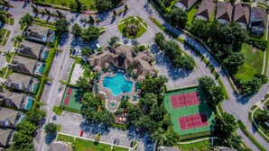 An aerial view of a residential area with a central swimming pool, tennis courts, and a landscaped park area.