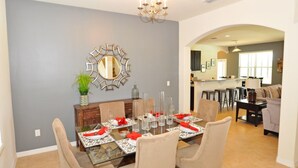Comedor con mesa de vidrio dispuesta para seis personas, rodeado de sillas color beige. Un espejo decorativo y una planta adornan la pared.