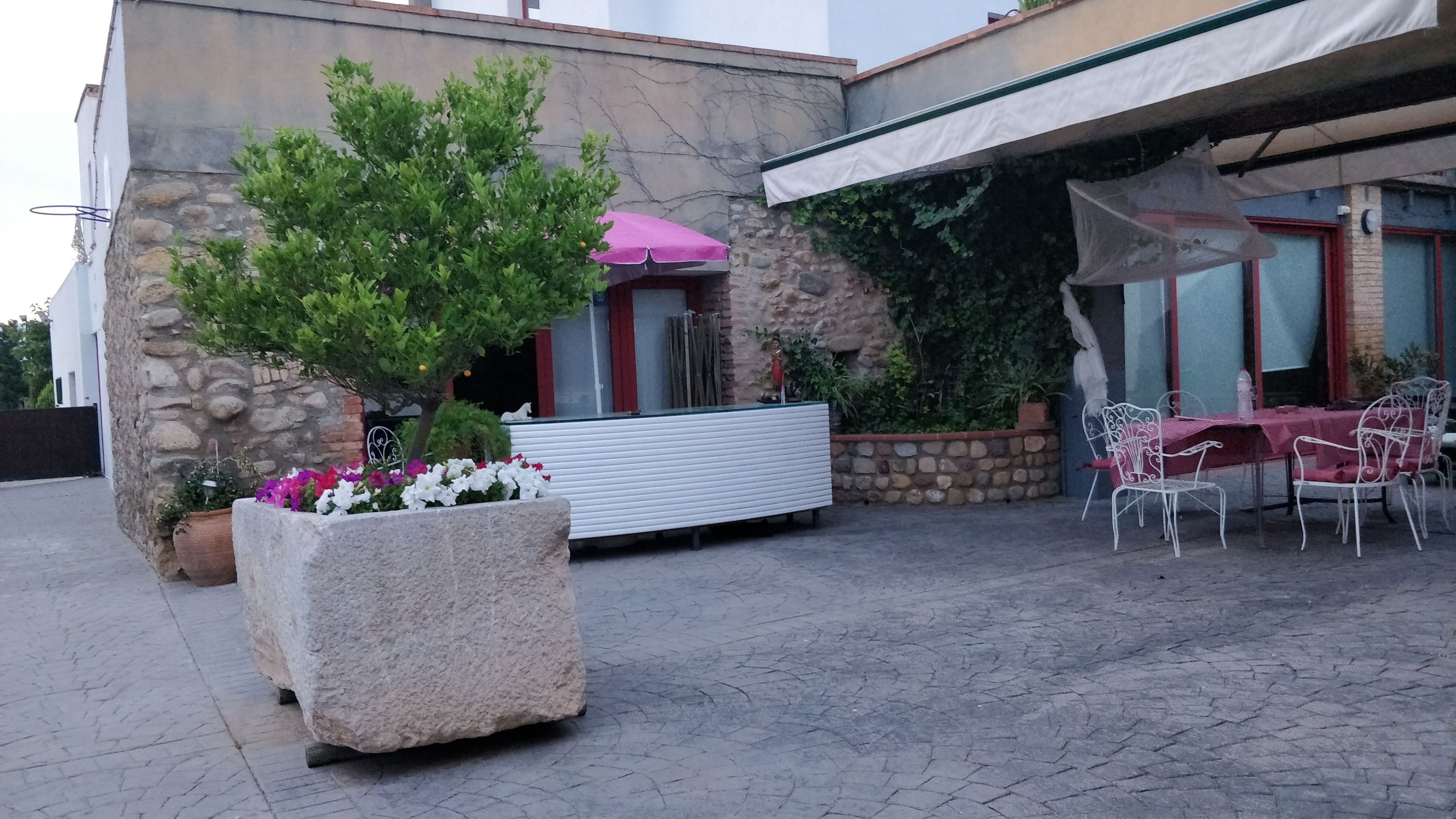 A stone building with a patio area featuring a white planter with flowers, a pink umbrella, and outdoor seating with white metal chairs and a table.