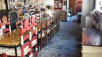 Un comedor con una larga mesa de madera y sillas, suelo de piedra, y diversos objetos en estanterías y paredes.
