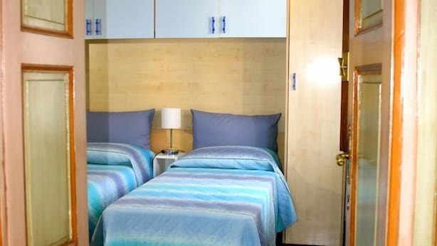 A hotel room with a wooden floor, two beds with blue bedding, a bedside table with a lamp, and light-colored walls. - Ai Cappuccini (Racconigi)