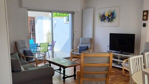 Un salon comprenant un canapé, une chaise en bois, un téléviseur et une table. Une porte-fenêtre coulissante donne accès à un espace extérieur aménagé avec une table et des chaises.