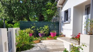 Un chemin dallé de pierres mène à un bâtiment blanc, bordé d’une clôture verte et agrémenté de plantes en pot.