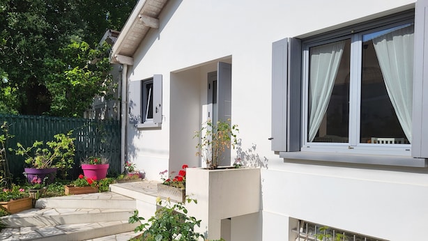 Une maison moderne à l’extérieur blanc, avec un chemin de pierre menant à l’entrée, et une variété de plantes et de fleurs en pot disposées autour de la propriété.