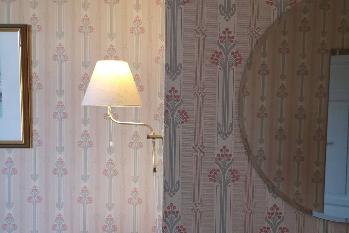 A bedroom with floral wallpaper, a bed with white bedding, a wall-mounted lamp, and a round mirror.