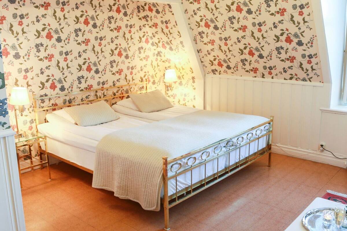 A bedroom with a bed, floral wallpaper, a radiator, and a bedside table with a lamp.