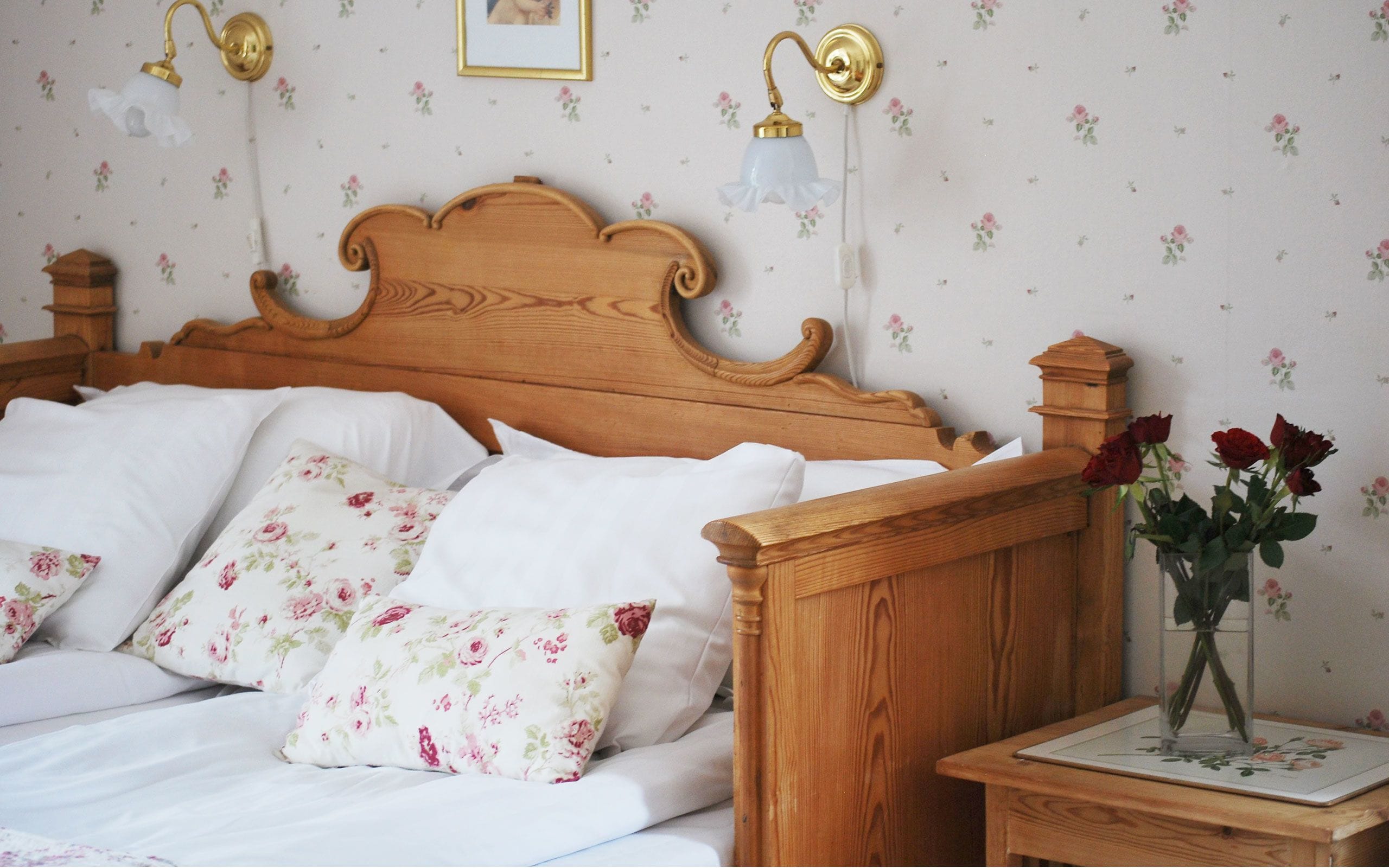 Una cama de madera con almohadas blancas y florales, una mesita de noche con un jarrón de rosas y lámparas fijadas a la pared.