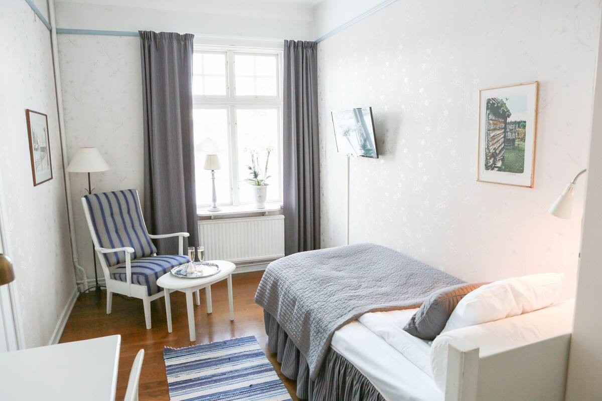 A bedroom with a bed, a chair, a small table, a lamp, a window with curtains, and a wall-mounted TV.