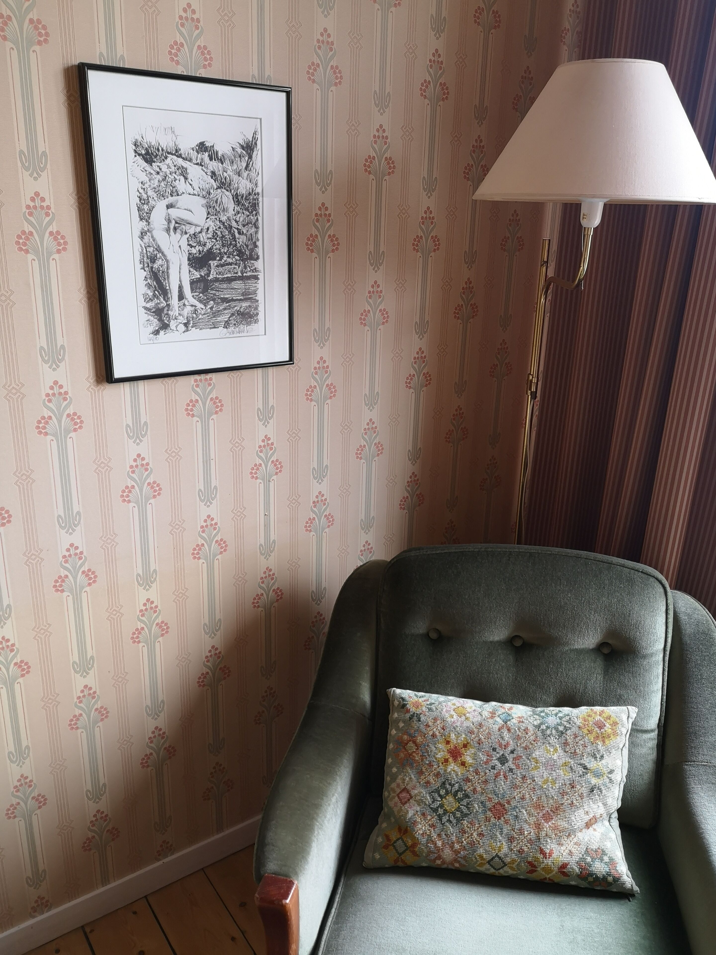 A room with a patterned wallpaper, a framed black and white artwork, a grey armchair with a colorful cushion, and a floor lamp.