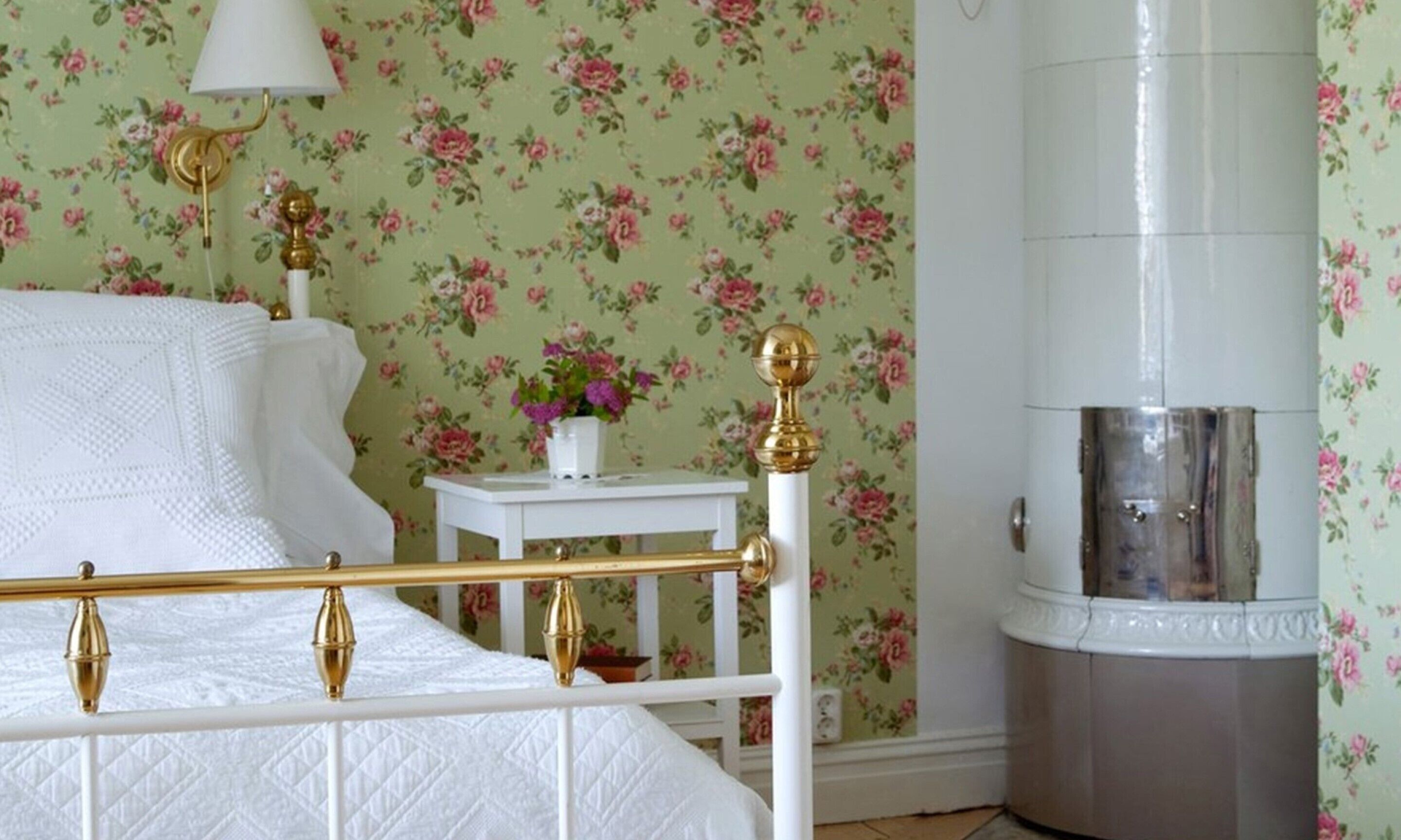 Une chambre avec un lit blanc, un papier peint à motifs floraux, une table de chevet blanche ornée d’un vase de fleurs et une colonne blanche avec une base métallique.