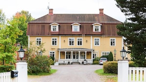 A two-story building with a symmetrical facade, a central entrance with a porch, and a gravel driveway leading to the entrance. - Korstäppans Herrgård (Leksand)