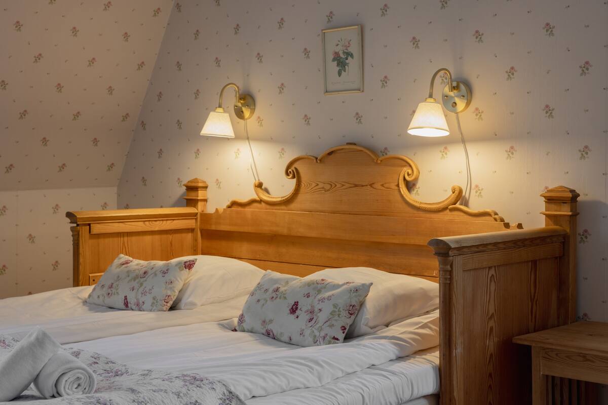 A neatly made bed with floral pillows and a wooden headboard, two wall-mounted lamps, and a framed picture on the wall.