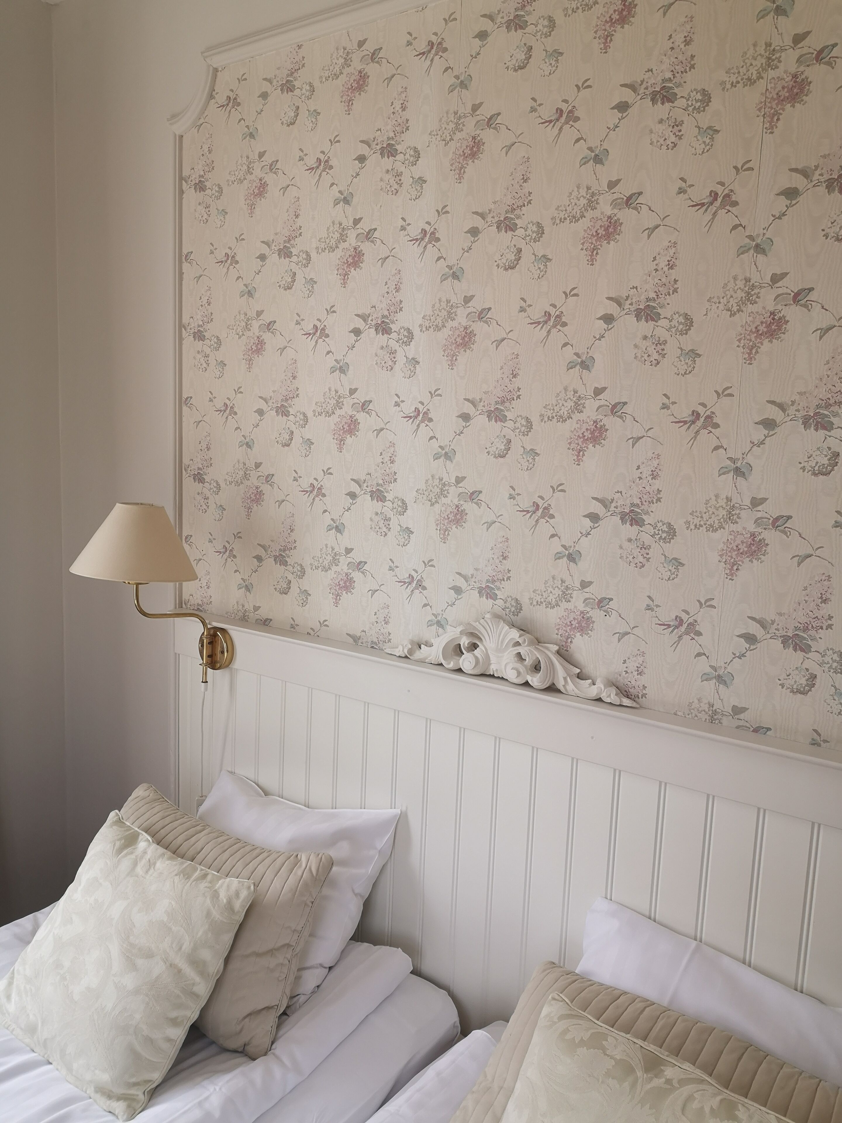 Un dormitorio con papel tapiz floral, una lámpara de noche y almohadas ordenadas.
