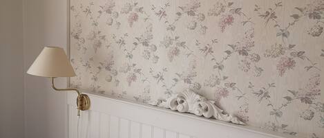 Un dormitorio con papel tapiz floral, una lámpara de noche y almohadas ordenadas.
