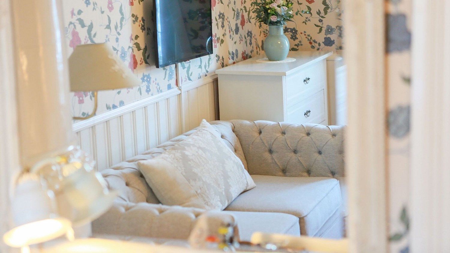 A bedroom with a tufted sofa, a bedside table with a vase, and floral wallpaper.
