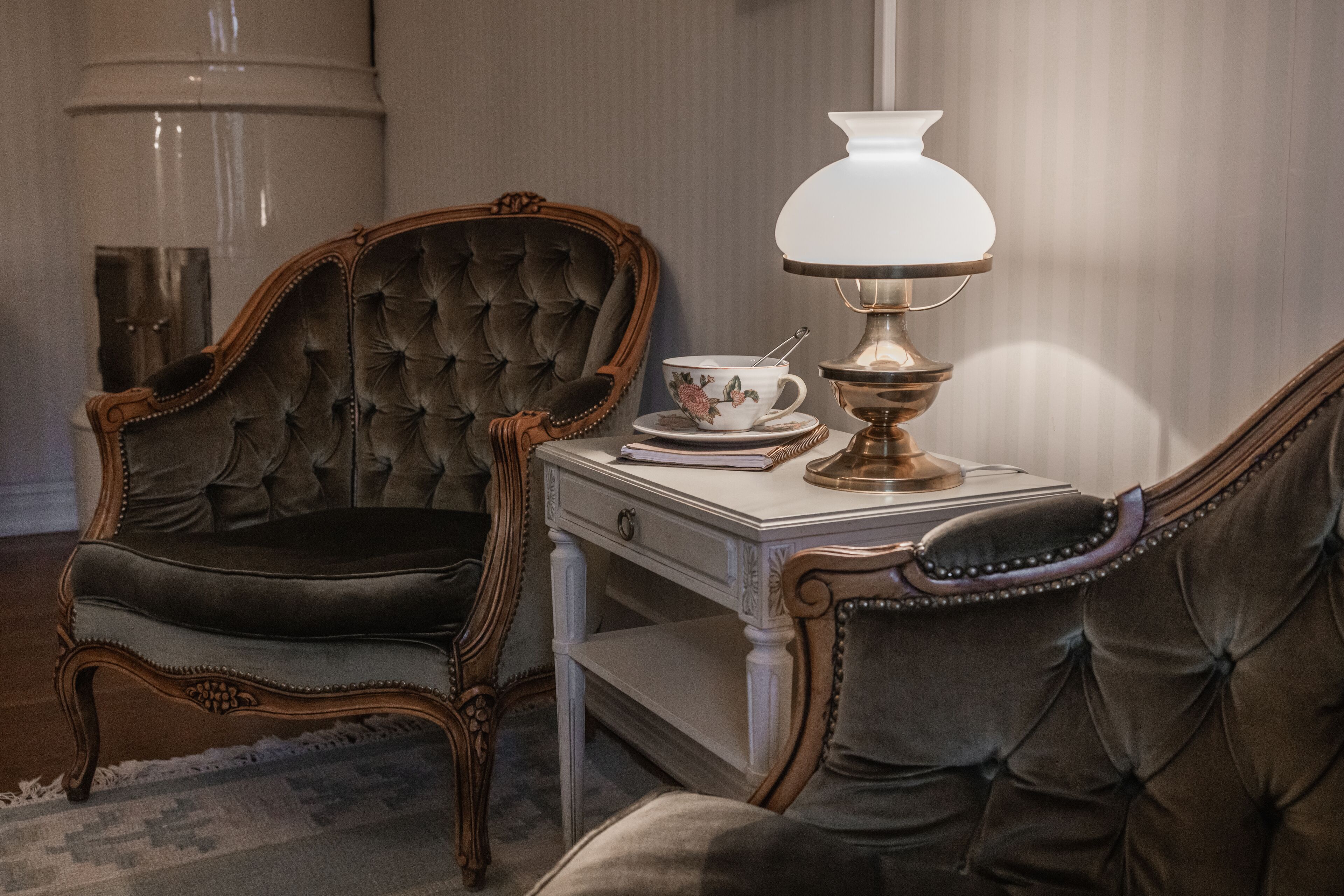 A room with a tufted leather armchair, a matching sofa, a white side table with a lamp, and a decorative plate.