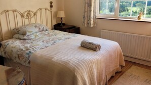 Un dormitorio con cama, piso de madera, ventana con vista a follaje y una lámpara, además de un cuadro en la pared.