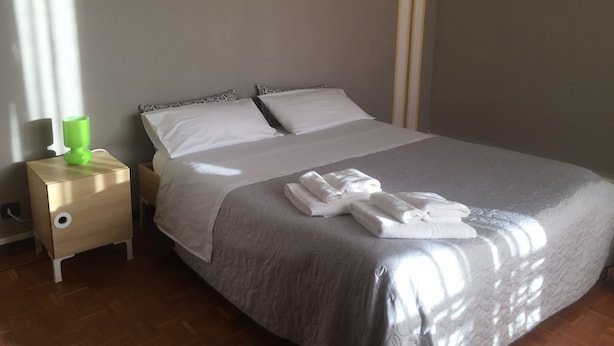 A neatly made bed with white linens and a grey quilt, a bedside table with a green lamp, and a wooden floor. - Casa Salice (Racconigi)